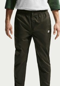 Homme portant un pantalon Nike noir avec une taille élastique et un bouton, mains détendues le long du corps, taille visible jusqu’aux cuisses inférieures.