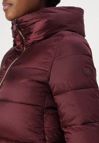 Burgundska puffer jakna s visokim ovratnikom, zatvaranjem na patentni zatvarač i kviltanom teksturom. Sadrži logotip na lijevom ramenu. Glatka površina tkanine.