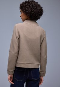 Vrouw met krullend haar, gekleed in een gestructureerd beige jack en donkerblauwe jeans, zijwaarts gekeerd tegen een effen grijze achtergrond.