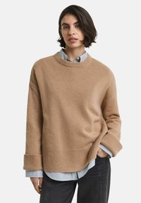 Pull en tricot beige avec un col rond, des épaules tombantes et des poignets côtelés, superposé à une chemise rayée bleue et blanche.
