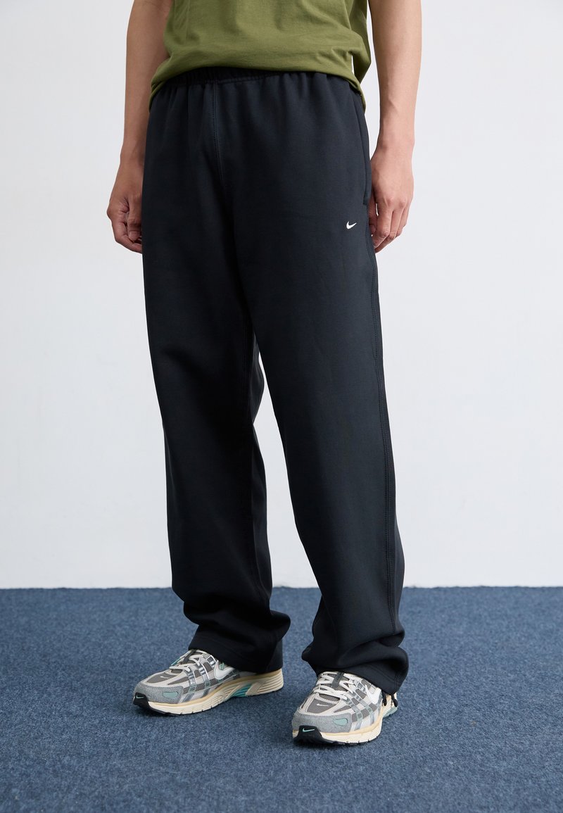 Schwarze Sporthose mit elastischem Bund, seitlichen Nähten und kleinem weißem Nike-Logo, kombiniert mit grauen und beigen stilisierten Laufschuhen.