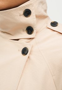 Γιακάς μπεζ καπαρντίνας σφικτά κλειστός με τρία σκουρόχρωμα κουμπιά απόχερσα χελώνα, κοντινή λήψη υφής υφάσματος και λεπτομερειών ραφής.