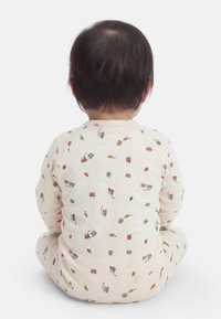 Βρέφος με σκούρα μαλλιά καθιστό σε σταυροπόδι, φορώντας ανοιχτόχρωμες πιτζάμες με μικρά πολύχρωμα σχέδια οχημάτων.