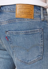 Levi's 502 farmer nadrág világoskék denim színben, barna bőr címkével, két hátsó zsebbel és a bal zseben található jellegzetes piros címkével.