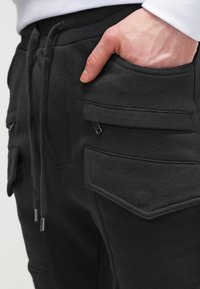 Černé joggingové kalhoty s texturovanou tkaninou, bočními kapsami se zipy a nastavitelnými šňůrkami v pase. Lehce a volně střižený design.