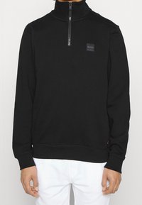 Sudadera negra con cremallera de cuarto y un pequeño logo "BOSS" en el pecho, combinada con pantalones blancos, mostrada desde el cuello hasta las caderas.