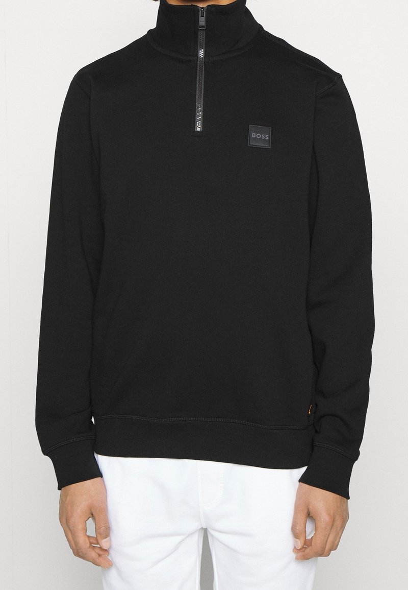 Sudadera negra con cremallera de cuarto y un pequeño logo "BOSS" en el pecho, combinada con pantalones blancos, mostrada desde el cuello hasta las caderas.
