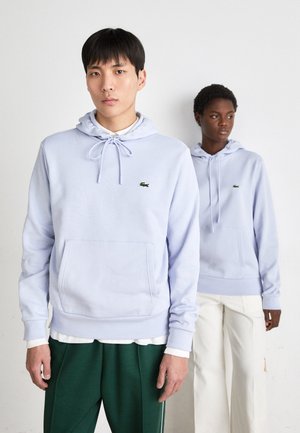 Sudaderas de lavanda claro con cordones, bolsillo frontal y pequeño logo verde. Un modelo lleva pantalones verdes y otro lleva pantalones blancos.
