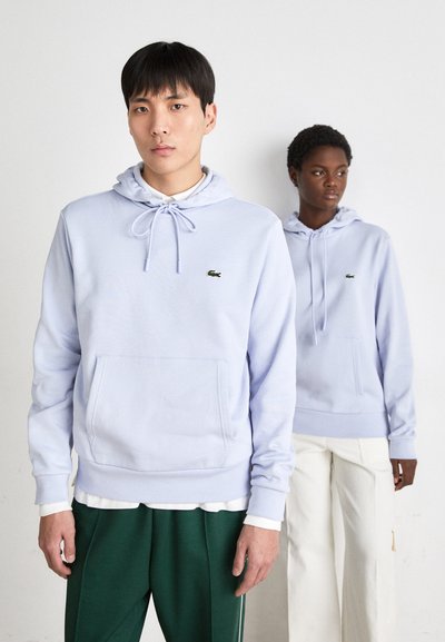 Hoodies lavande clair avec cordons, poche avant et petit logo vert. Un mannequin porte un pantalon vert, l'autre un pantalon blanc.