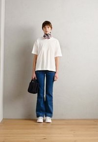 Vit t-shirt med kort ärm, kombinerad med utsvängda blåjeans, vita sneakers och en svart handväska. Har en mönstrad halsduk runt nacken.