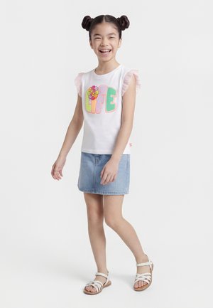 Fille souriante aux cheveux en deux chignons, portant un haut blanc à manches volantées avec le mot "LIFE" et un motif floral, une jupe en jean et des sandales blanches, debout.