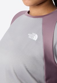 Maglietta atletica grigia con tessuto strutturato, maniche raglan viola e un logo bianco. Il colletto è rotondo e aderente.