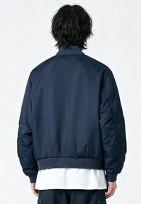 Giacca bomber blu navy realizzata in tessuto liscio; presenta un colletto e polsini a costine, orlo elastico e una vestibilità rilassata, con cuciture laterali visibili.