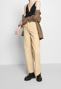 Pantalones anchos beige combinados con una camisola de encaje negra y una camisa grande de cuadros marrones, con botas de tobillo negras y gruesas.