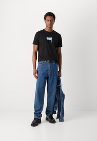 Camiseta de algodón negra con un estampado gráfico azul, combinada con unos vaqueros de denim azules sueltos y zapatos de plataforma negros, sosteniendo una chaqueta de denim.