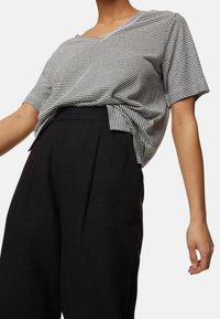 Personne portant un haut ample à manches courtes rayé noir et blanc, rentré dans un pantalon noir ample taille haute sur fond blanc.