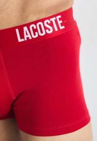 Red boxer briefs with a white elastic waistband featuring the "LACOSTE" logo, made from smooth, stretchable fabric.