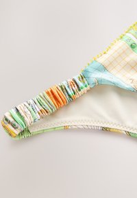 Lähikuva bikinialushousujen kulmasta, jossa on moniväriset joustavat sivukoristeet ja vaalealle kankaalle painettu karttaa muistuttava kuvio.