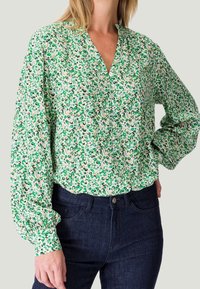 Blusa verde y blanca con estampado, de mangas largas, escote en V y corte relajado, combinada con jeans de mezclilla azul oscuro.