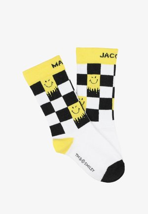 Chaussettes à carreaux noir et blanc avec des accents jaunes et un motif de smiley. Poignets côtes ; sections noire pour les orteils et le talon. Matériau en mélange de coton.