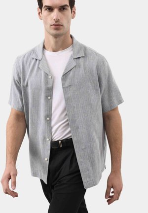 Mężczyzna ubrany w jasnoszarą koszulę z krótkim rękawem na białym t-shircie oraz czarne spodnie, idzie do przodu na jednolitym tle.