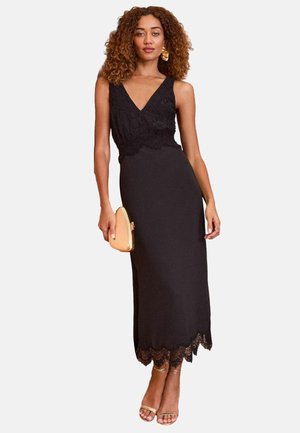 Abito lungo nero con bustier in pizzo, scollo a V e orlo in pizzo ondulato. L'abito è abbinato a una borsa a tracolla dorata e sandali con cinturino.