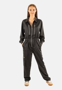 Jumpsuit noir zippé avec manches longues, taille élastique et deux poches zippées à l'avant. Fini mat, coupe décontractée, associé à des baskets.