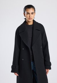 Dubbelknopsgemaakt zwart wollen jasje met grote ingesneden kraag, knopenaccenten en manchetten details. Model draagt het met een fitted top en jeans.