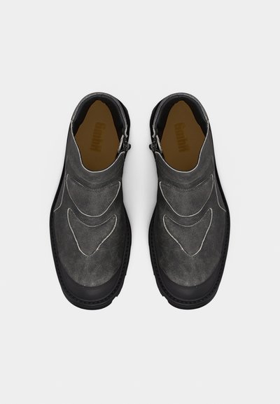 Ζευγάρι σκούρων γκρι σουεντ παπουτσιών τύπου slip-on με μαύρες σόλες και διακριτικό ραμμένο σχέδιο, απεικονίζεται από επάνω σε λευκό φόντο.