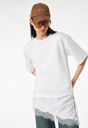 Femme portant un t-shirt blanc oversize avec un ourlet en dentelle, un jean bleu, une casquette marron "SELF LOVE" et des lunettes de soleil teintées orange sur un fond uni.