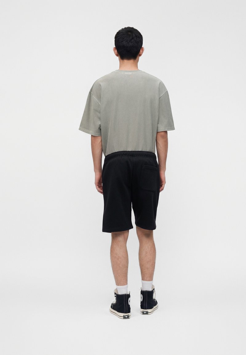 Junger Mann steht mit dem Rücken zur Kamera, trägt ein hellgraues oversized T-Shirt, schwarze knielange Shorts, weiße Socken und schwarze High-Top-Sneaker.