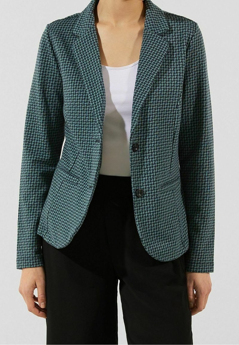 Femme portant un blazer à motifs verts et noirs sur un haut blanc et un pantalon noir, debout devant un fond uni.