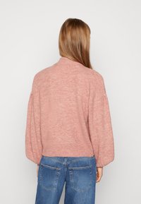 Ljusrosa stickad tröja med hög krage, sänkta axlar och lös passform. Bärs över blå denimjeans, som visar rundad nederkant och struktur.