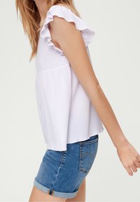 Widok z boku osoby ubrane w biały, marszczony top bez rękawów i podwinięte szorty dżinsowe na jednolitym jasnym tle.