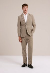 Homme portant un costume beige, une chemise blanche et des chaussures de ville noires devant un fond beige uni.