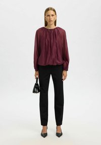 Viininpunainen, pitkähihainen paita kerätyllä kaula-aukolla, yhdistettynä mustiin, istuviin housuihin ja mustiin, kärjestä teräviin korkokenkiin, pitäen pientä käsilaukkua.