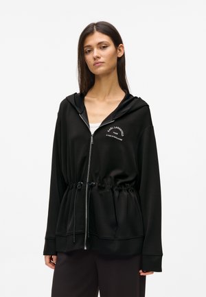 Jeune femme portant un sweat à capuche noir zippé avec une taille à cordon de serrage et un logo texte blanc sur la poitrine, debout devant un fond uni.