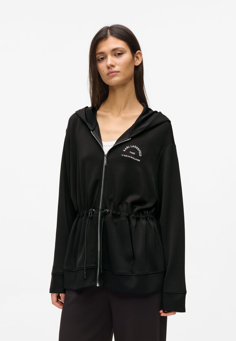 Νεαρή γυναίκα που φοράει μαύρο φούτερ με φερμουάρ και κορδόνι στη μέση, με λευκό κείμενο λογότυπο στο στήθος, στέκεται μπροστά σε ένα απλό φόντο.