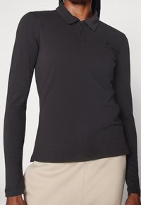 Person som bär en figurnära svart långärmad polo med knäppning och beige byxor mot en enfärgad ljus bakgrund.