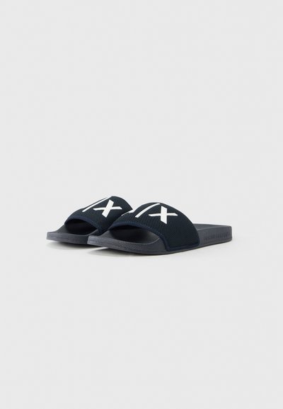 Sandales à glissière bleu marine avec dessus en mesh, présentant un motif croisé blanc. Semelle plate en caoutchouc, style à bout ouvert, et forme minimaliste.