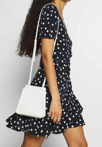 Žena v tmavě modrých šatech s puntíky a volány, nese malou bílou crossbody kabelku s texturovaným vzorem.