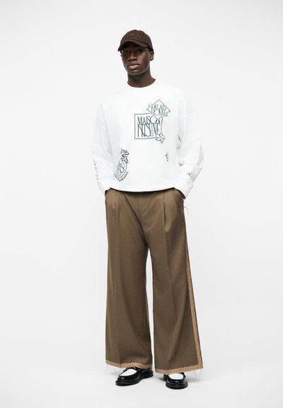 Άντρας που φοράει ένα λευκό φούτερ με σχέδιο, καφέ παντελόνι φαρδιάς γραμμής με λωρίδα στο πλάι, μαύρο καπέλο, λευκές κάλτσες και μαύρα παπούτσια, στέκεται με τα χέρια στις τσέπες.