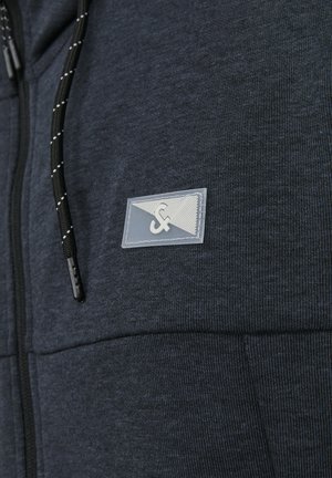 Navy blue zip-up hoodie with textured fabric, featuring a clear logo patch and black drawstring with white accents.