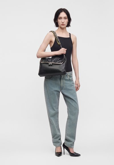 Femme en débardeur noir, jean bleu clair et talons noirs tenant un grand sac à main noir avec une bandoulière chaîne sur un fond uni.