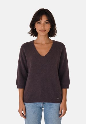 Mujer con cabello corto y oscuro que lleva un suéter de cuello en V de color púrpura oscuro con mangas tres cuartos y jeans azul claro, de pie frente a un fondo liso.