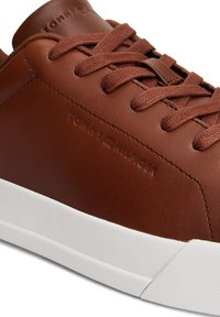 Nahaufnahme eines braunen Ledersneakers mit passenden Schnürsenkeln, weißer Sohle und geprägtem Tommy Hilfiger-Logo an der Seite und der Zunge.