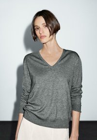 Een vrouw met kort bruin haar draagt een grijze V-hals sweater, kijkt recht vooruit tegen een minimalistische achtergrond en straalt een kalme uitdrukking uit.