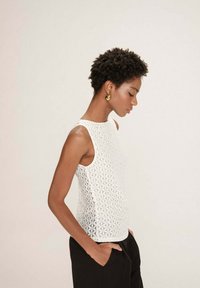 Femme aux cheveux courts et bouclés portant des boucles d'oreilles en forme de goutte d'or, un haut sans manches à motifs blancs et un pantalon noir, les mains dans les poches.