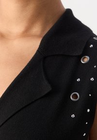 Gros plan sur un vêtement noir sans manches avec un col plié, présentant des œillets métalliques et de petites clous brillants sur la zone des épaules.
