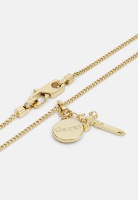 Gouden ketting met een karabijnsluiting, een ronde hanger met de inscriptie "Guess" en een kruispendant als detail.